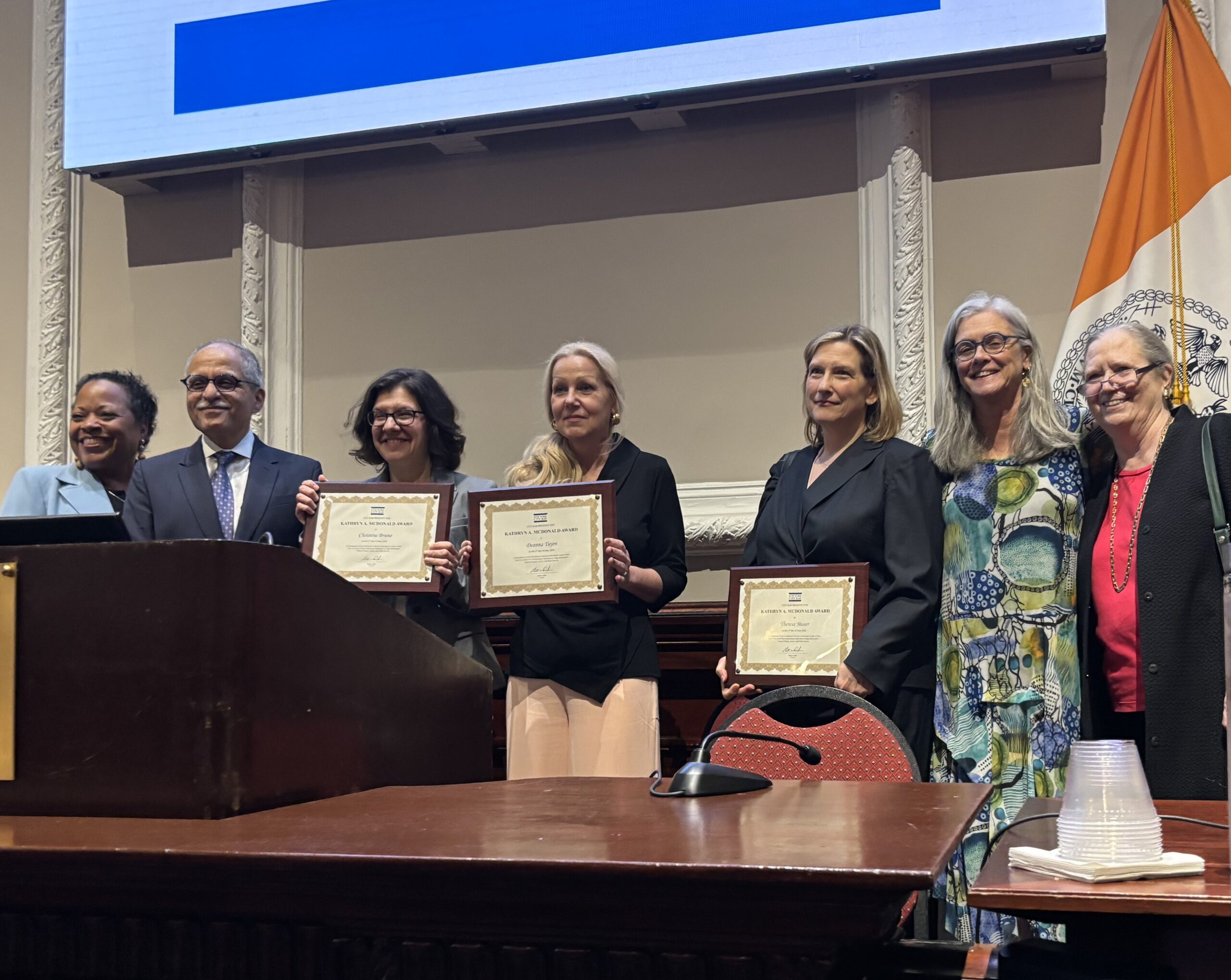 A group of people posing in front of a camera. Three women are holding up awards.